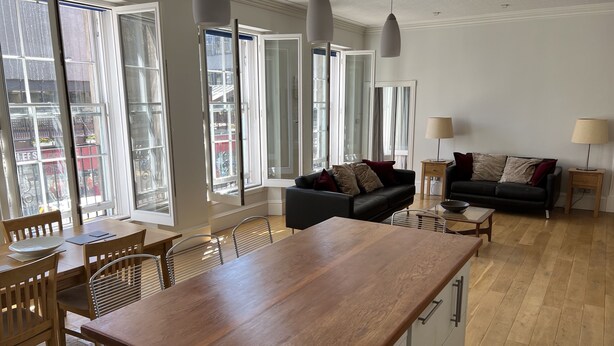 Un vaste espace de vie comprenant un coin repas avec une table en bois, des canapés noirs et un coin cuisine doté d’un plan de travail en bois.