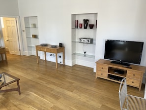 Uma sala de estar moderna com piso de madeira, mesa de centro de vidro, aparador para TV com tela plana, estante com objetos decorativos e porta que dá para outro cômodo.