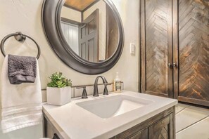 Un baño con un lavamanos blanco, un espejo redondo, un perchero para toallas, una planta en maceta y una puerta de madera.