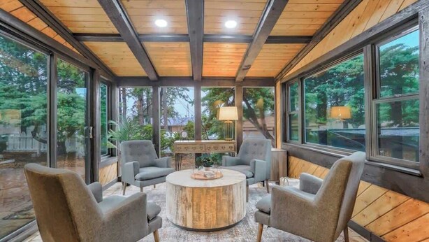 Un interior moderno con una mesa de centro redonda de madera, rodeada por cuatro sillones grises, ventanas amplias que ofrecen vistas al exterior y un techo con vigas de madera a la vista.