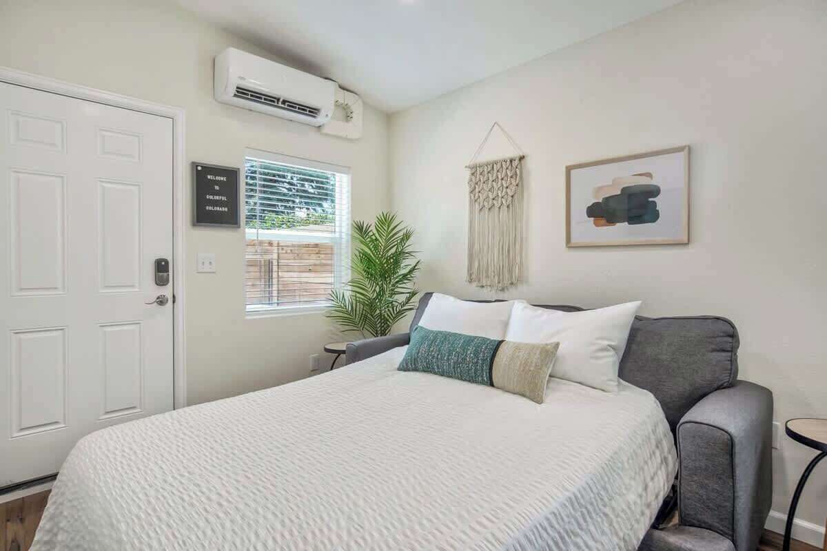 Une chambre à coucher avec un lit, un fauteuil gris, une décoration murale en macramé et un tableau encadré.