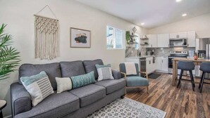 A modern living room with a grey sofa, a coffee table, and a patterned rug. The kitchen is visible in the background with white cabinets and stainless steel appliances.