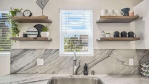 A kitchen with marble countertops, a sink, and wooden shelves with kitchenware.