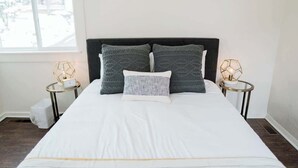 A neatly made bed with a white and gray striped bedspread, two matching pillows, and a dark headboard. There are two bedside tables with lamps and a window with a view of trees.