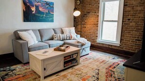 A living room with a gray sectional sofa, a white coffee table, a colorful rug, a large window, and a brick wall.