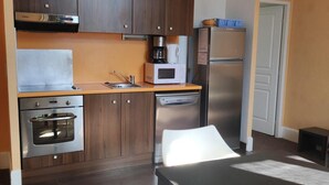 Ett kompakt kök med mörka träskåp, en ugn i rostfritt stål, mikrovågsugn och kylskåp. Det finns ett litet matbord med två stolar.