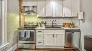A compact kitchen with white cabinets, a brick wall, a stainless steel oven, and a dishwasher. - Miners Escape! Timeless District Cozy Haven (Colorado Springs)