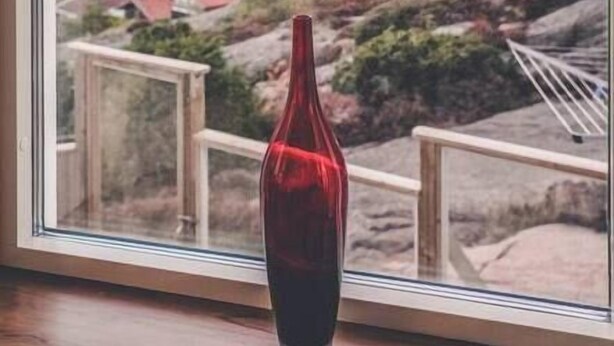 Ein Fenster mit Blick auf einen Sonnenuntergang über einer Küstenlandschaft, eine rote Flasche im Vordergrund und ein hölzerner Geländer.