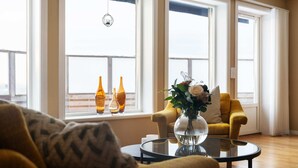 A living room with a yellow armchair, a glass coffee table with a vase, and a window with three bottles.