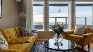 A living room with a yellow sofa, a round glass coffee table, and a yellow armchair. There are two windows with curtains, a hanging light fixture, and a vase with flowers on the coffee table.