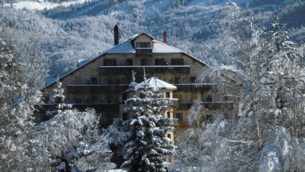 Un edificio grande con varios balcones, rodeado de árboles nevados y montañas al fondo.