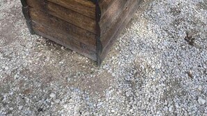 Una jardinera de madera con una variedad de flores y plantas, ubicada sobre una superficie de grava.