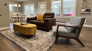 Una sala de estar con un sofá de cuero marrón, un reposapiés amarillo y un sillón gris. Se observa una alfombra estampada en el suelo, y en el fondo, un comedor con sillas blancas y una mesa.