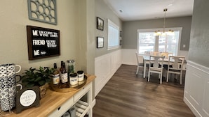 Un comedor con mesa de madera, sillas y una lámpara de araña. Hay un letrero en la pared con un mensaje divertido.