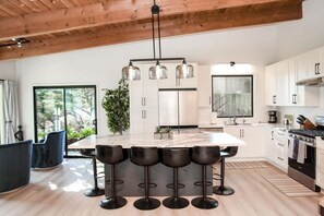 Una cocina moderna con una isla central, taburetes de bar, armarios blancos y un ventanal grande.