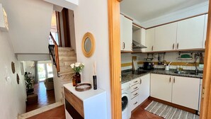 Eine kleine Küche mit weißen Schränken, einem eingebauten Ofen und einem Spülbecken. Eine Holztreppe führt zu einer anderen Ebene, und an der Wand hängt ein Spiegel.