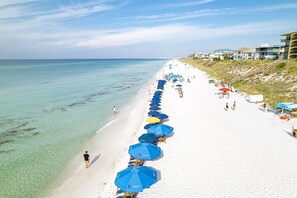 A beach with rows of blue umbrellas, clear turquoise water, and buildings on the right.