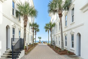 A pathway flanked by white buildings with palm trees, leading to a beach.