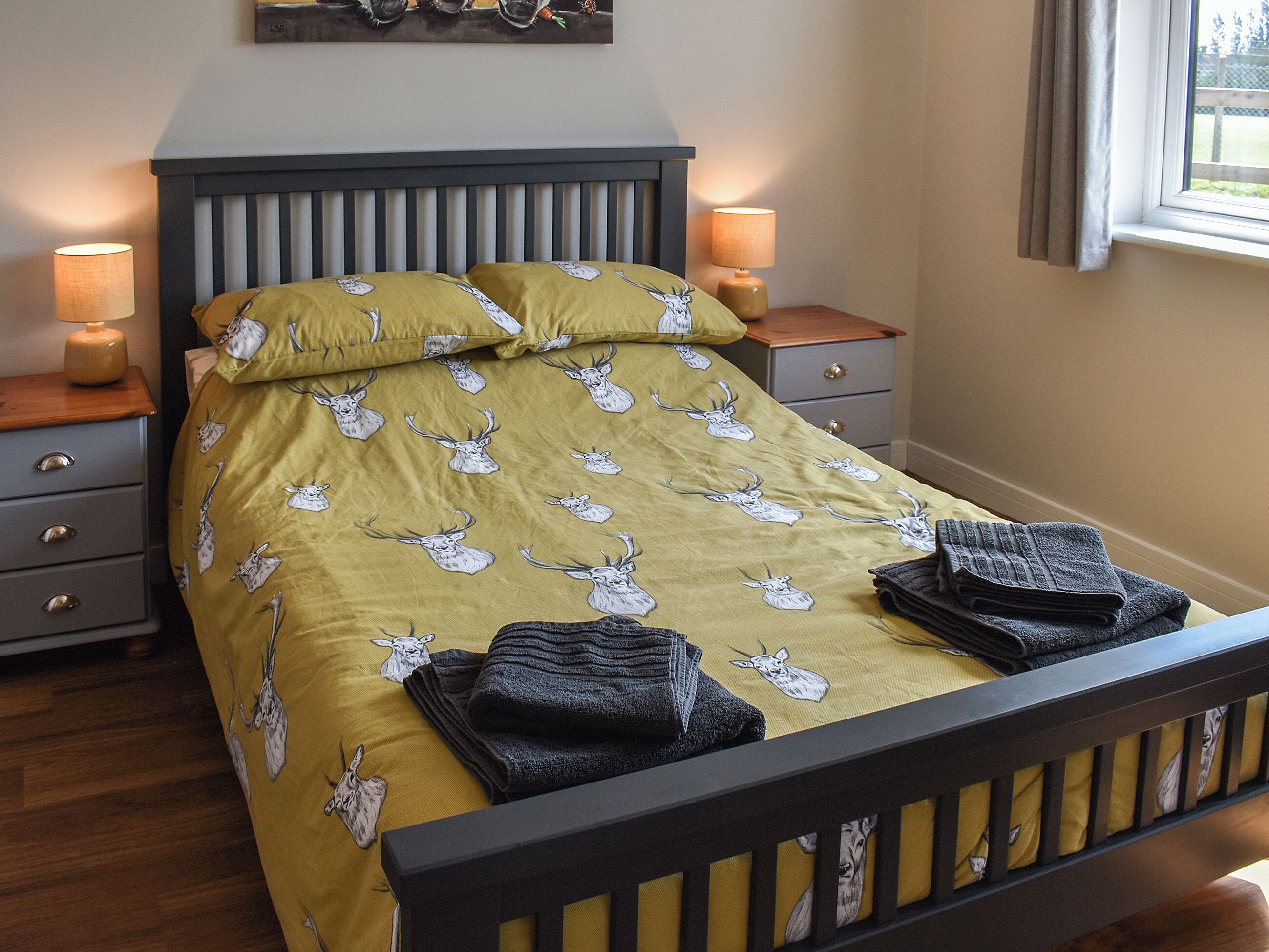 Een net opgemaakt bed met een beddekleed met patroon, twee bedlampjes, een nachtkastje en een raam met gordijnen.