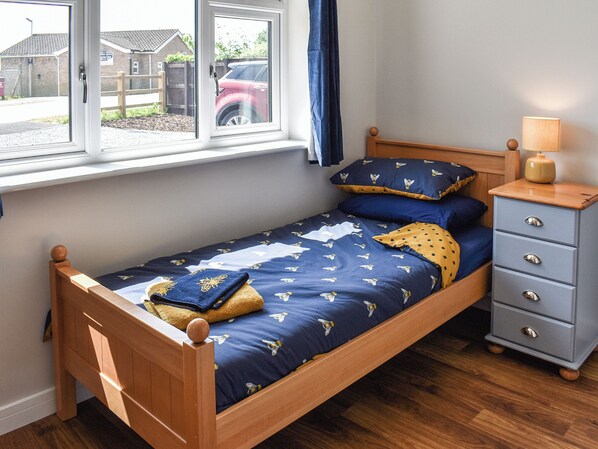 A single bed with a wooden frame and a blue bedspread with a bee pattern. There is a wooden nightstand with drawers and a lamp on it. A window with a view of a red car and a fence is visible. - 3 bedroom accommodation in Martham (Martham)