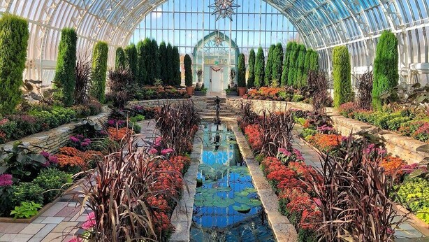Une serre dotée d’un toit en verre, d’une fontaine centrale et de diverses plantes et fleurs.