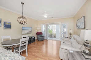 A spacious living room with a dining area, a gray sofa, a chandelier, and a large window with blinds.