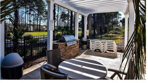 Een overdekt terras met een barbecue, buitenmeubels en uitzicht op een sportveld.