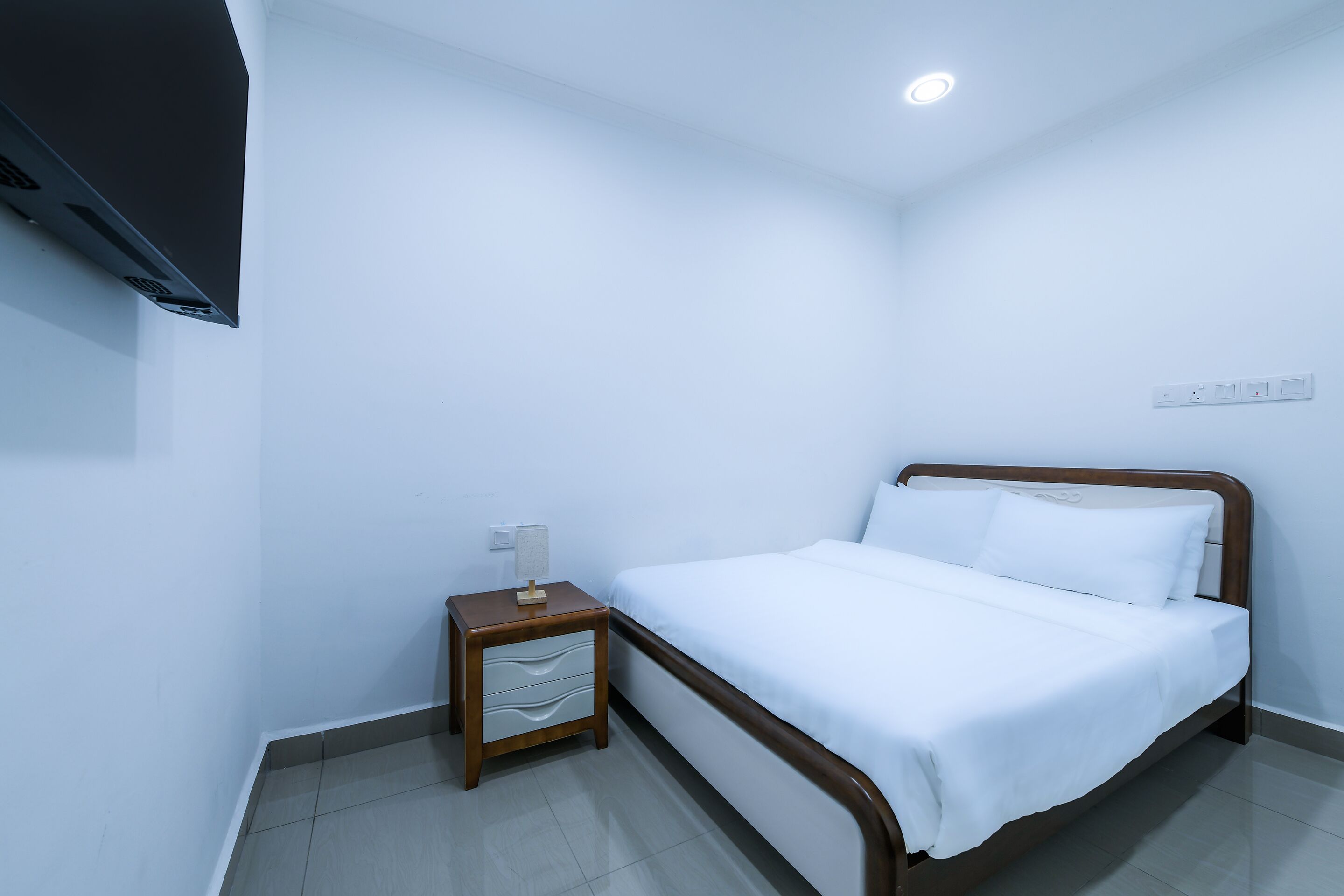 Una habitación de hotel con una cama, una mesita de noche, un televisor montado en la pared y una lámpara de techo.