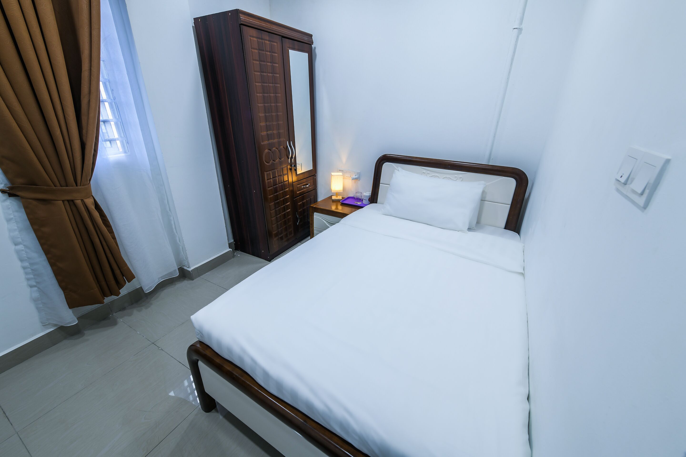 A hotel room with a single bed, a bedside table with a lamp, a wardrobe, and a curtain.