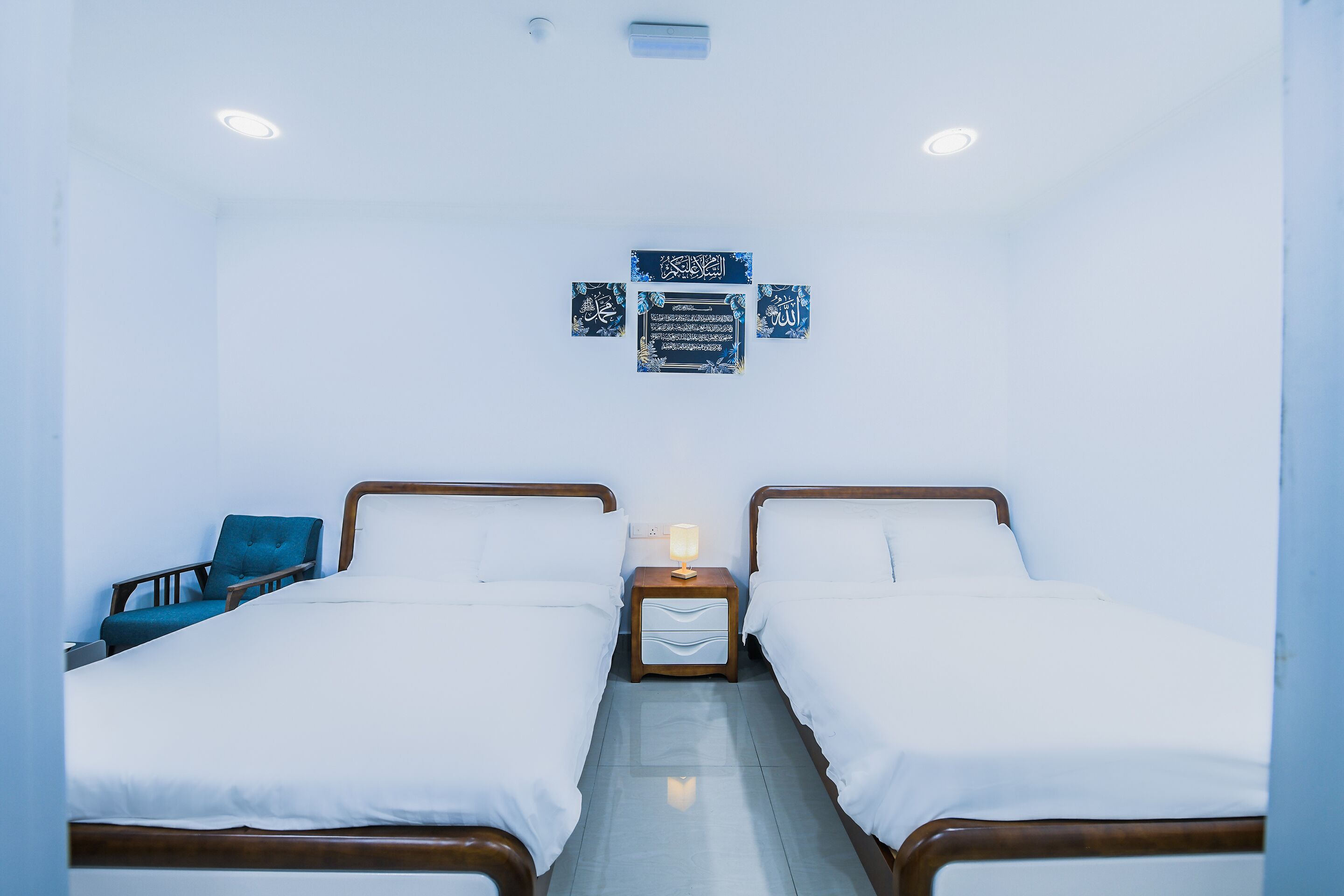A hotel room with two single beds, a nightstand with a lamp, a chair, and a wall-mounted decoration.