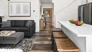 Ein modernes Wohnzimmer mit einem grauen Sofa, einem Couchtisch und einer Holzbank. Es gibt eine Küchenzeile mit einer weißen Arbeitsplatte und einem Kühlschrank.