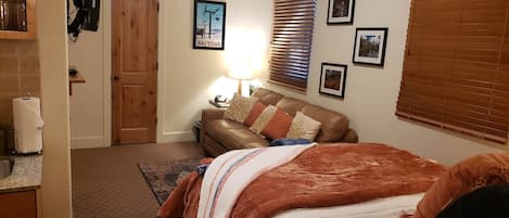 A bedroom with a bed, a wooden door, a brown leather couch, a lamp, and framed pictures on the wall.
