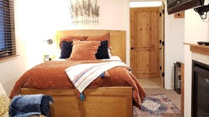 Un dormitorio con cama de madera, televisor montado en la pared, chimenea y puerta que da a otra habitación.