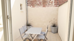 A small outdoor patio with a metal table and chairs, two potted plants, and a brick wall in the background.