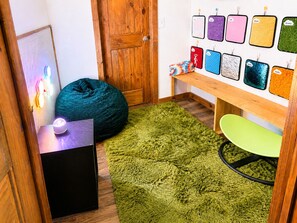Quarto com porta de madeira, tapete felpudo verde, um bean bag, uma mesa pequena com abajur e parede com diversos tapetes texturizados.