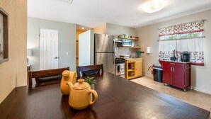 Cocina compacta con refrigerador, microondas, estufa y un lavamanos pequeño. Hay una mesa de comedor de madera con dos sillas y un banco. Se ve una ventana con cortinas, y una planta pequeña está sobre la mesa.