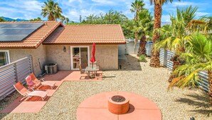 Una casa con techo de tejas, un patio con sillas y una mesa, un brasero y un jardín con palmeras.
