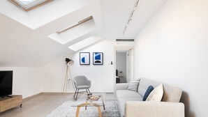 Ett modernt vardagsrum med högt tak, takfönster, en platt-TV, en grå fåtölj, en soffa med dekorativa kuddar och ett soffbord i glas.