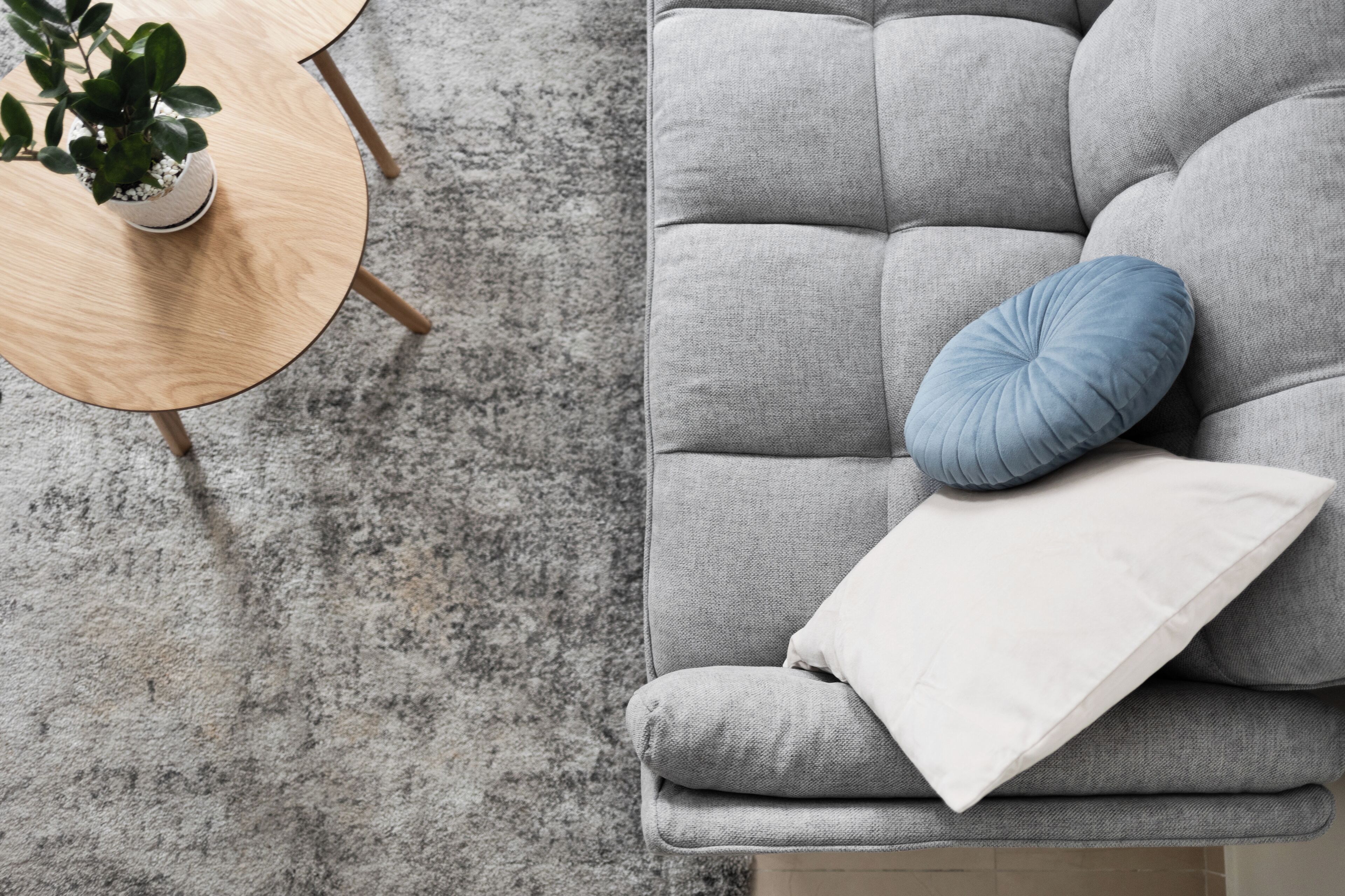 Un divano grigio con un cuscino bianco e blu, un piccolo tavolino con una pianta in vaso e un pavimento rivestito di moquette grigia.
