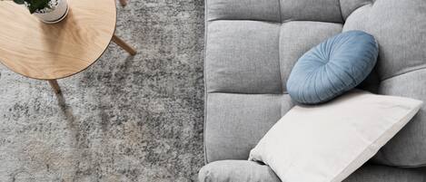 A grey sofa with a white and blue pillow, a small wooden table with a potted plant, and a grey carpeted floor.