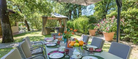 Un espacio de comedor al aire libre con una mesa preparada para cuatro personas, rodeada de sillas, bajo un toldo.