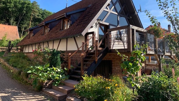 Et traditionelt hus med et stejlt tag, en trætrappe op til en balkon, og en have med forskellige planter og blomster.