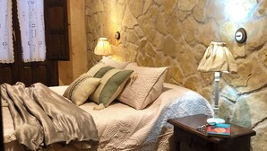 Une chambre avec un mur en pierre, un lit avec des oreillers, une table de chevet avec une lampe et une fenêtre avec des rideaux.
