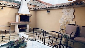 Une terrasse extérieure avec une table en verre, des chaises en métal, un mur en pierre et un four en briques.