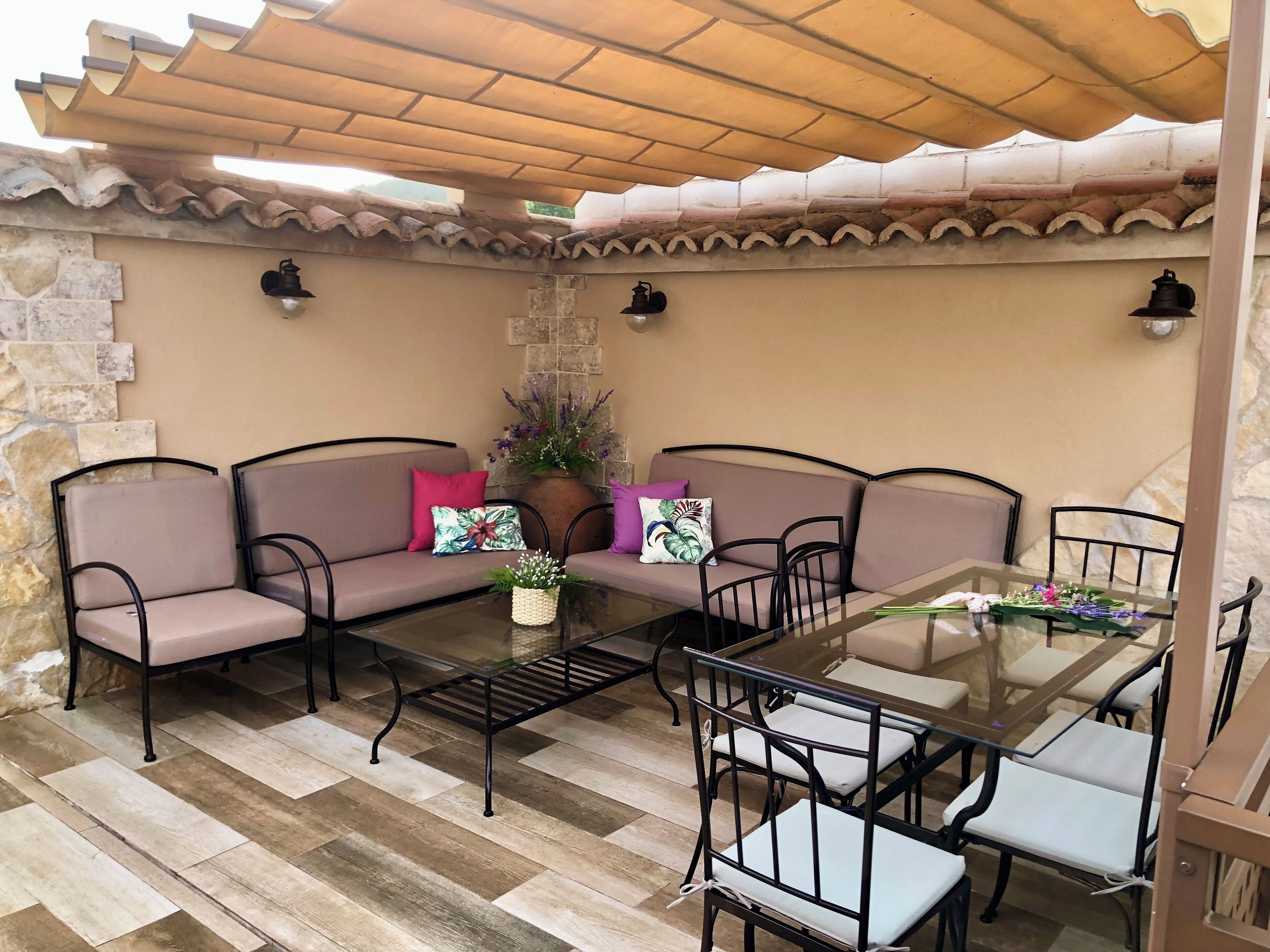 An outdoor patio with a stone wall, a wooden pergola, a cushioned sofa, a glass-top table, and metal chairs.