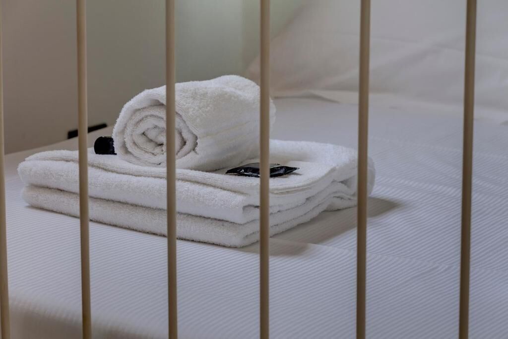 Una toalla blanca doblada cuidadosamente sobre una cama con un almohadón blanco y un objeto negro encima.