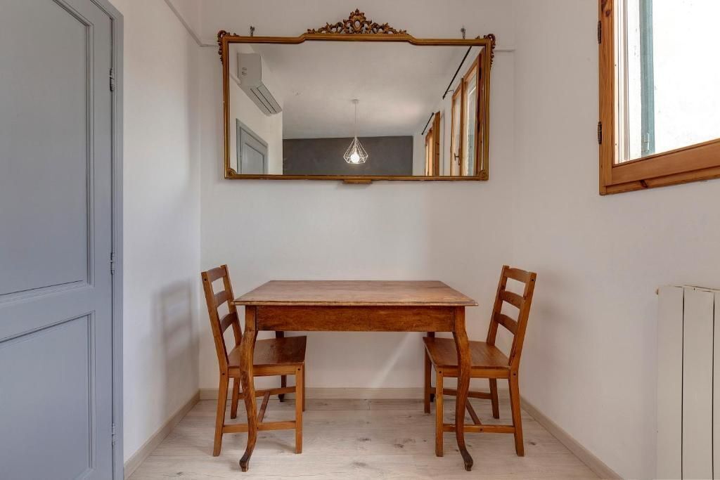 Uma sala de jantar com uma mesa e cadeiras de madeira, um espelho sobre a mesa e uma janela à direita.