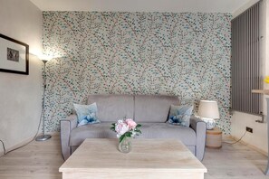 Uma sala de estar com um sofá cinzento, uma mesa de centro de madeira com um vaso de flores, um abajur e uma parede com um padrão de folhagens.