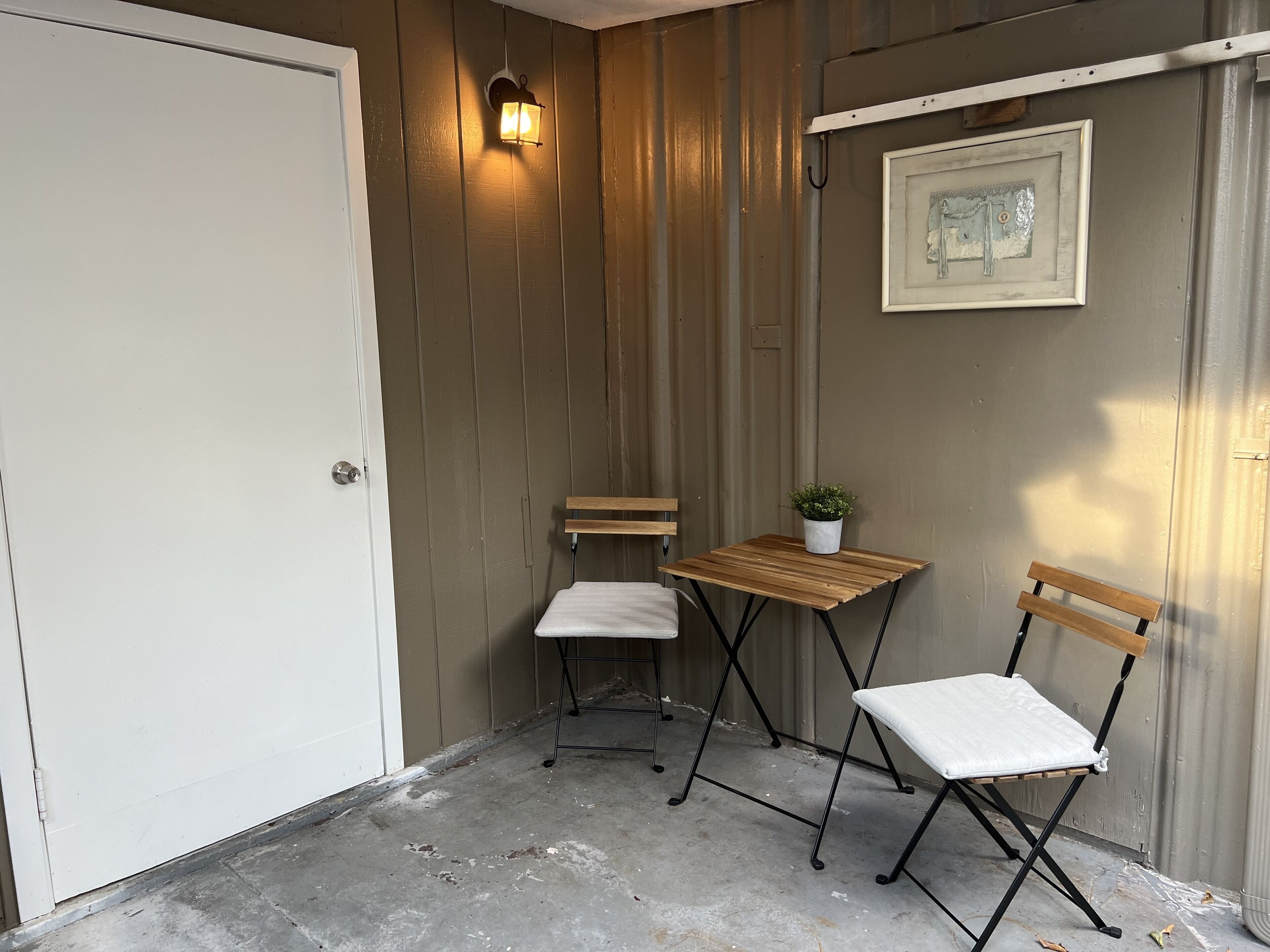 Ett litet, avgränsat utomhusområde med en dörr, två hopfällbara stolar, ett litet bord, en krukväxt och en väggmonterad lampa.