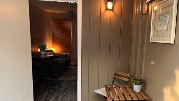 Un petit espace extérieur clos, aménagé avec un banc en bois et une table, un pot de fleurs et un luminaire fixé au mur.